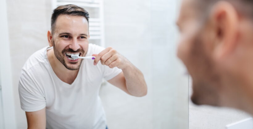 Handsome caucasian man standing in bathroom in pajamas and washing his teeth in the morning and watching himself in the mirror.