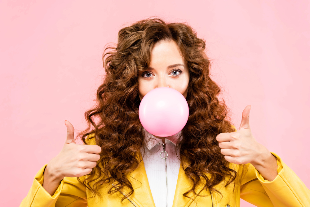 young woman with red, curly hair blowing a big bubble on pink background
