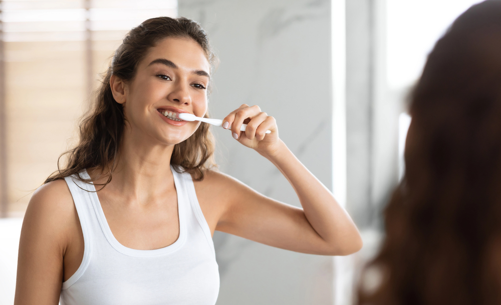 woman with brown curly hair and white tank top brushing her teeth, looking in mirror